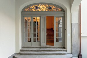 Un escalier en pierre mène à une porte à deux battants, dotée de panneaux de verre et surmontée d’une arche décorative.