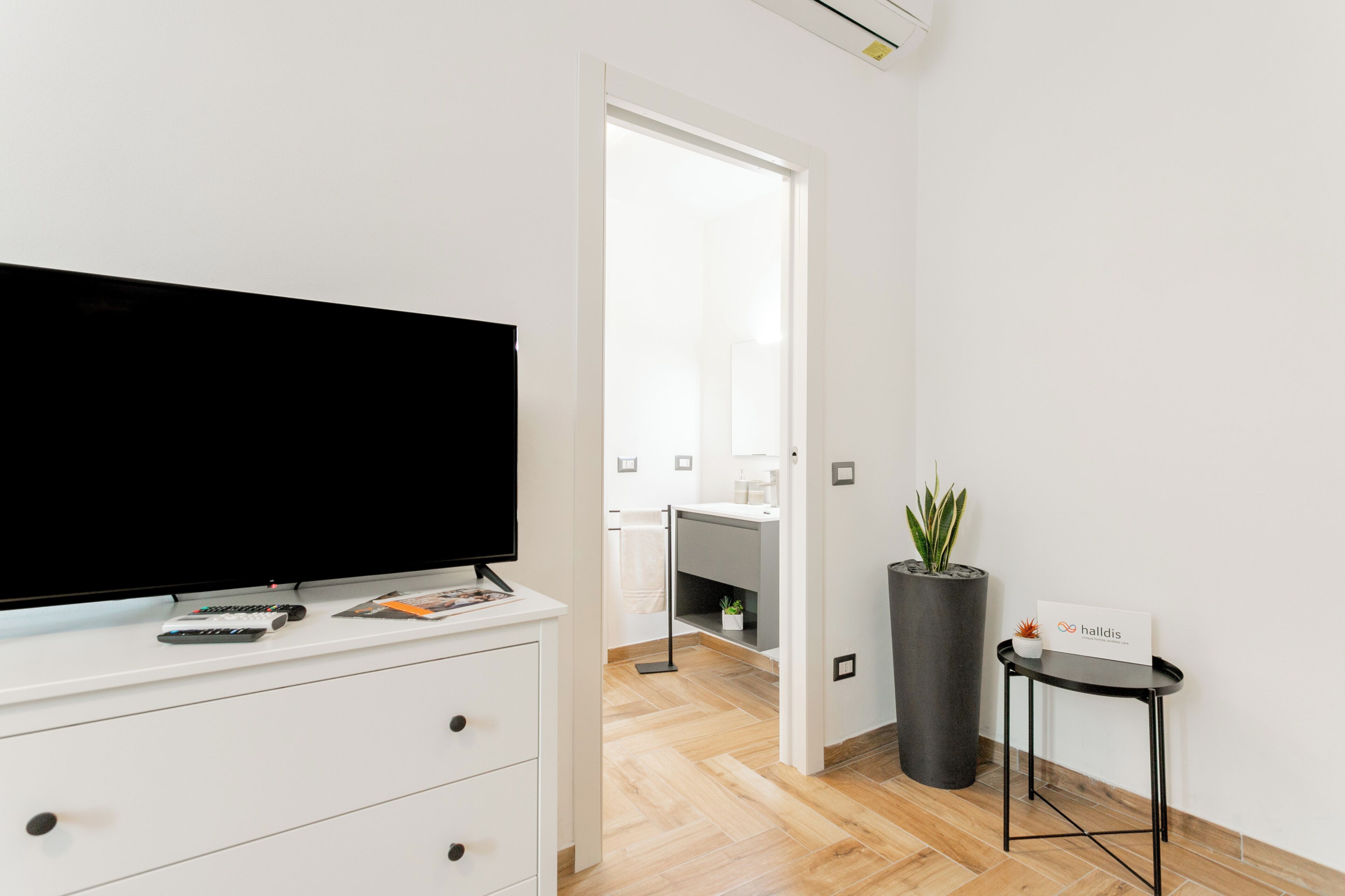 A modern room with a flat-screen TV on a white dresser, a potted plant, and a small table with a sign.