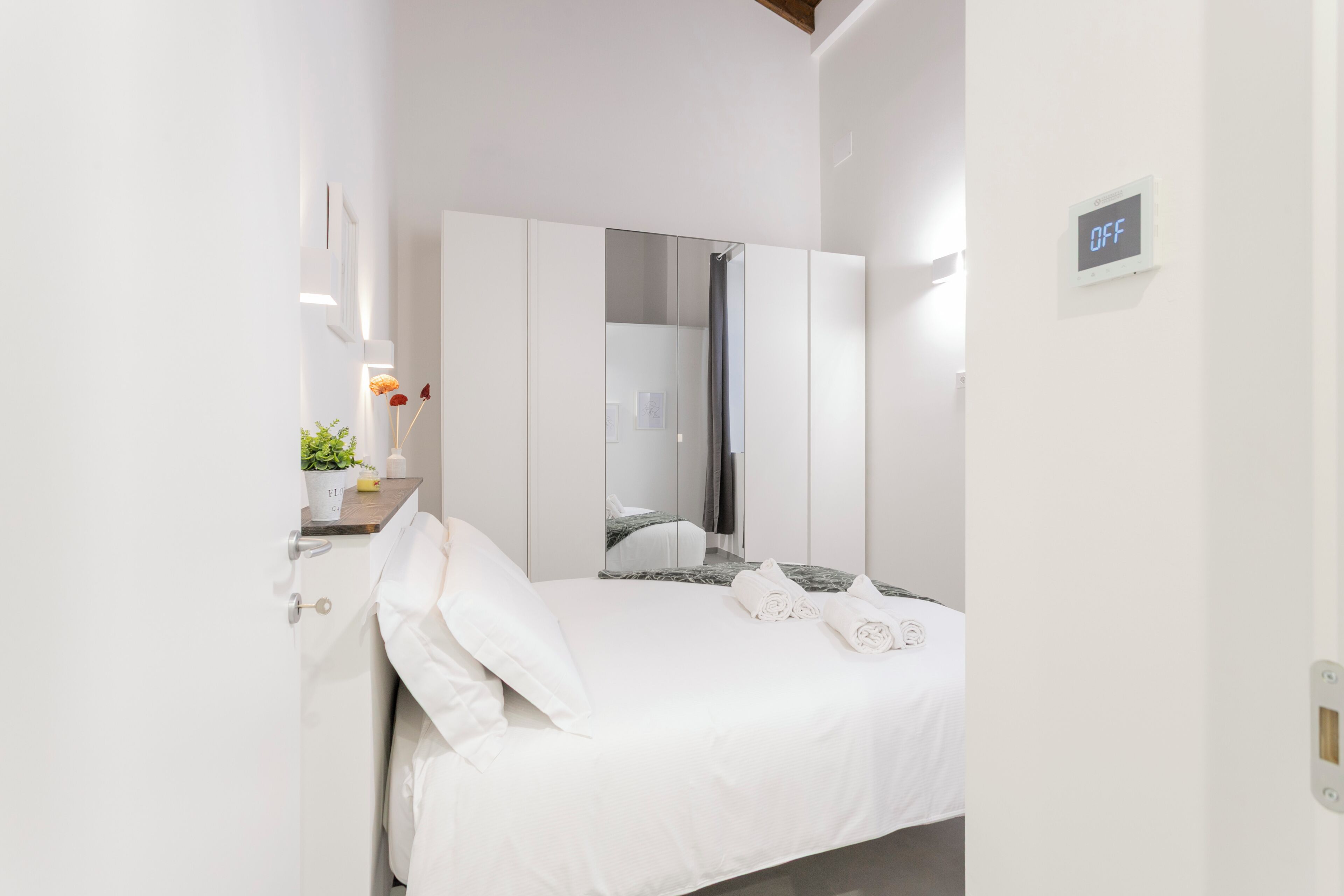 Une chambre d’hôtel moderne équipée d’un lit, d’un miroir, d’une armoire et d’une petite table agrémentée d’une plante.