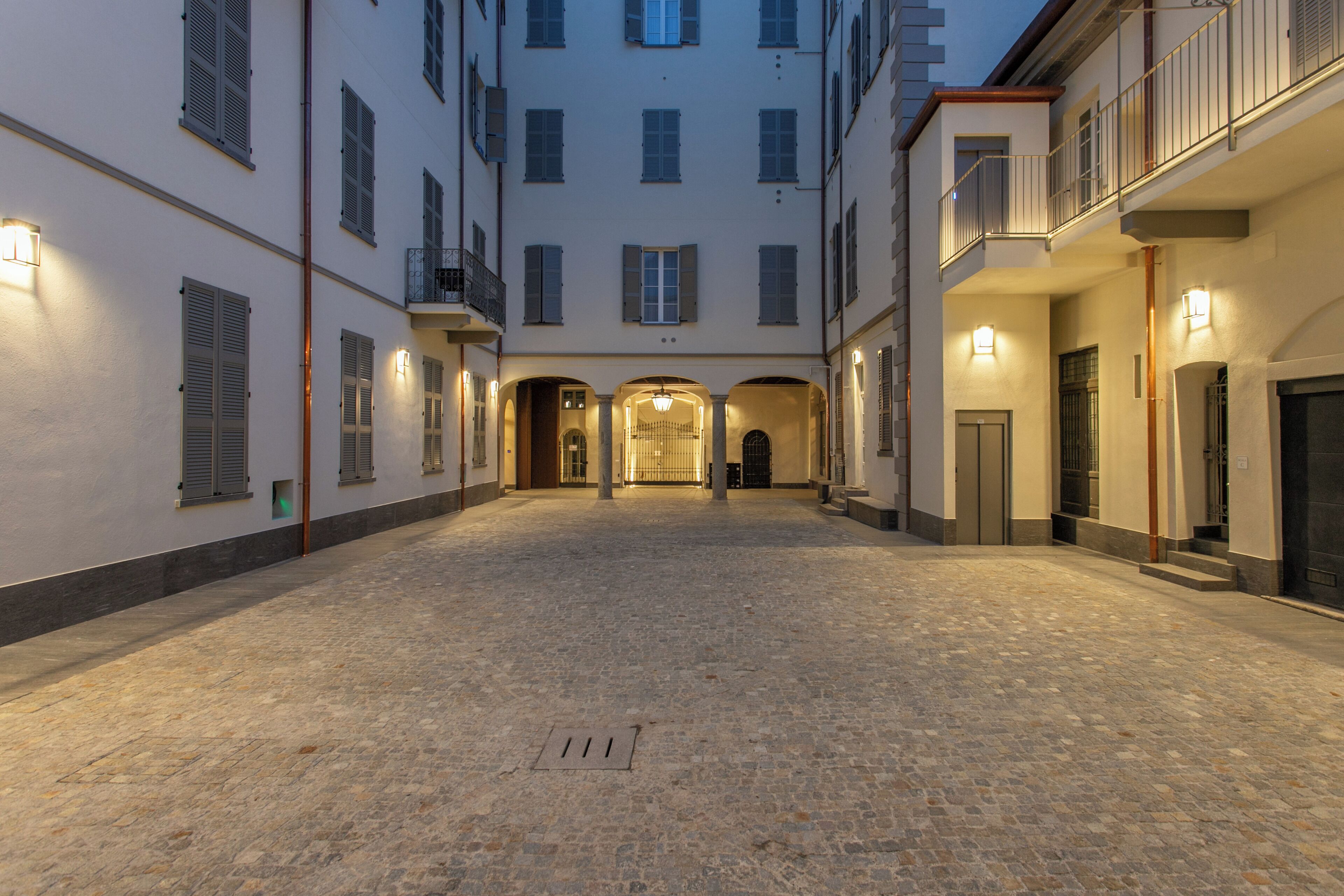 A courtyard with cobblestone flooring, multiple buildings with balconies, and illuminated exterior lights.