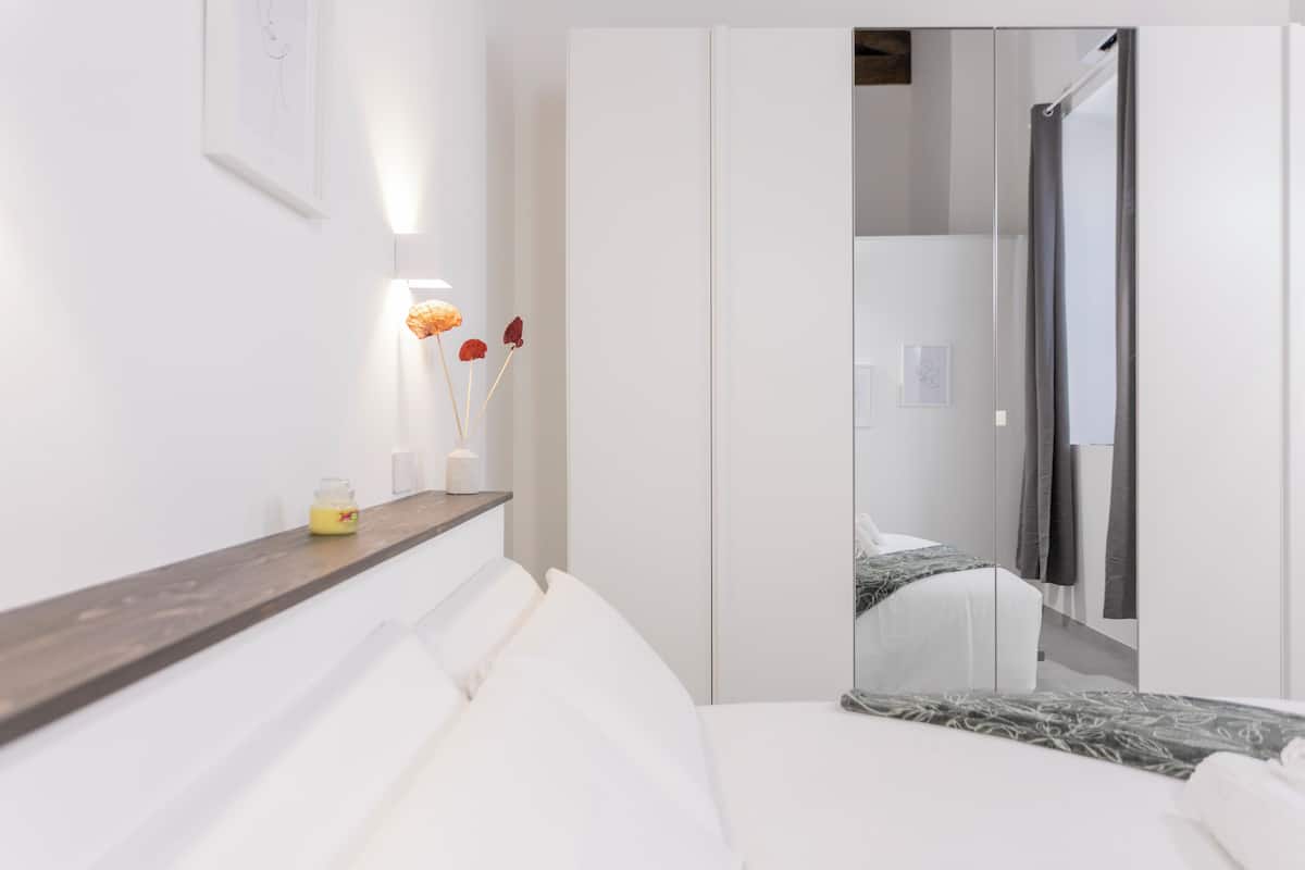 A modern hotel room with a bed, a bedside table with a vase of flowers, a mirror, and a wardrobe.