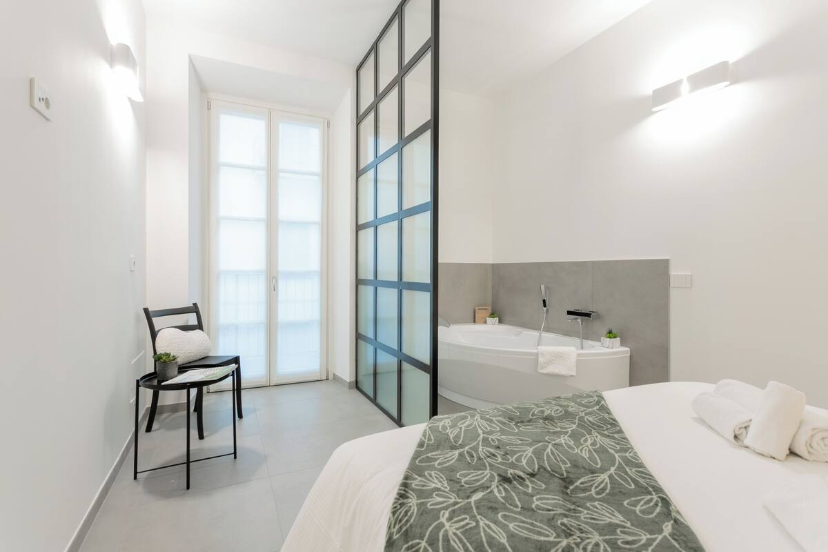 A modern hotel room with a bed, a chair, a large window, a bathtub, and a small plant.