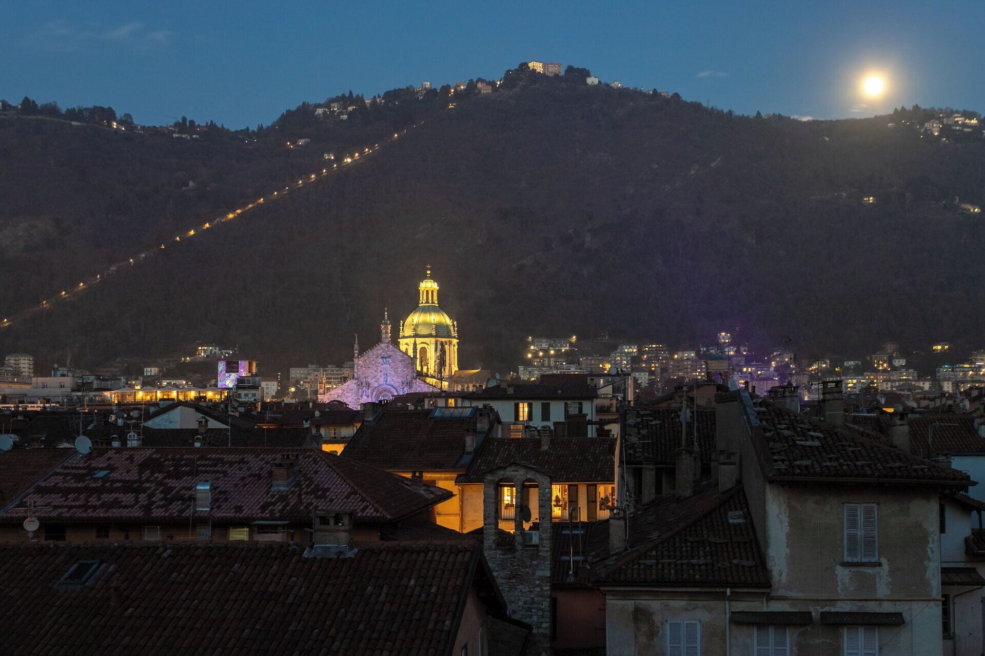 Une vue urbaine avec un dôme d’église illuminé qui se détache, une montagne en arrière-plan et un ciel nocturne dégagé.