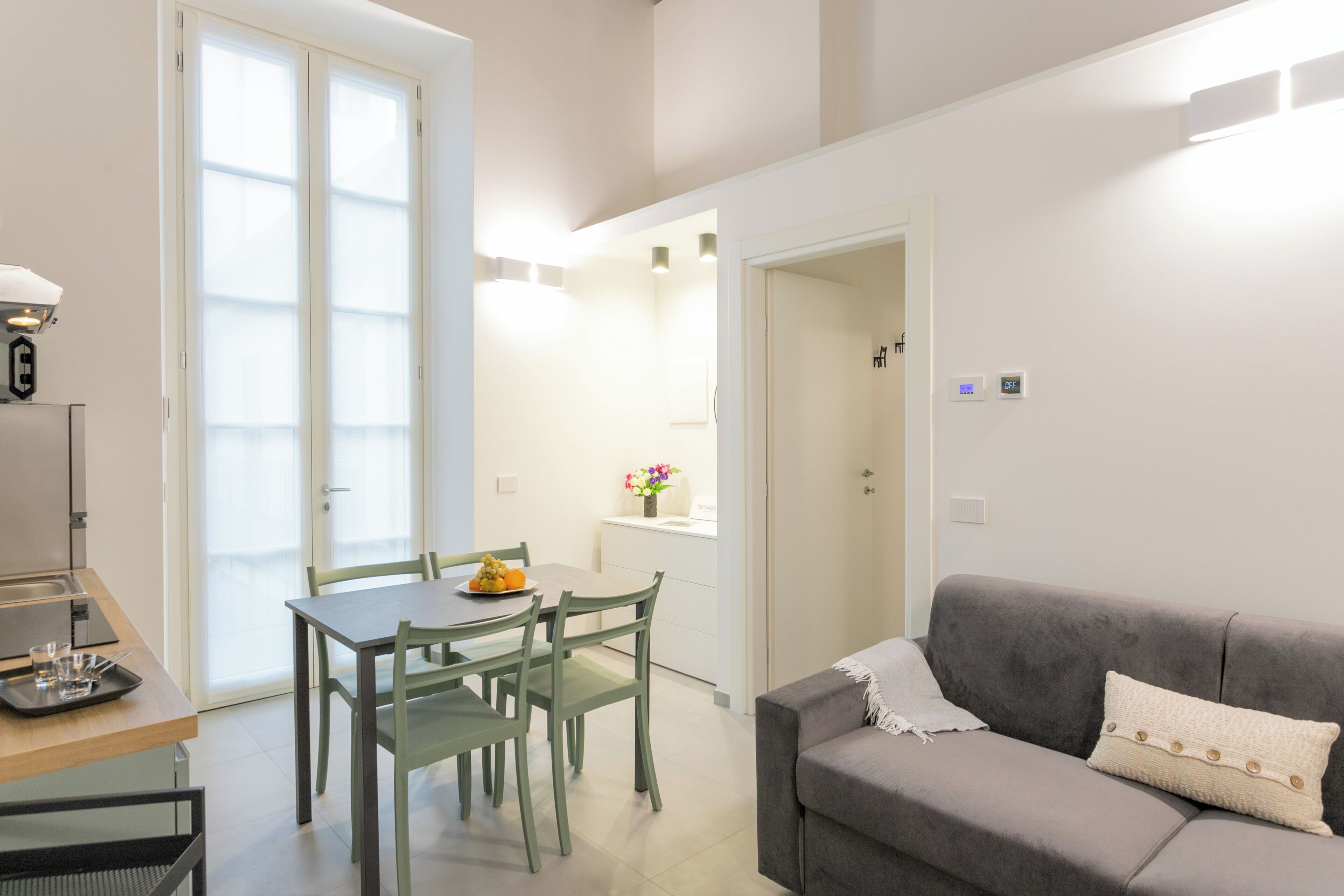 A modern living room with a grey sofa, a dining table with chairs, and a kitchen area with a window.