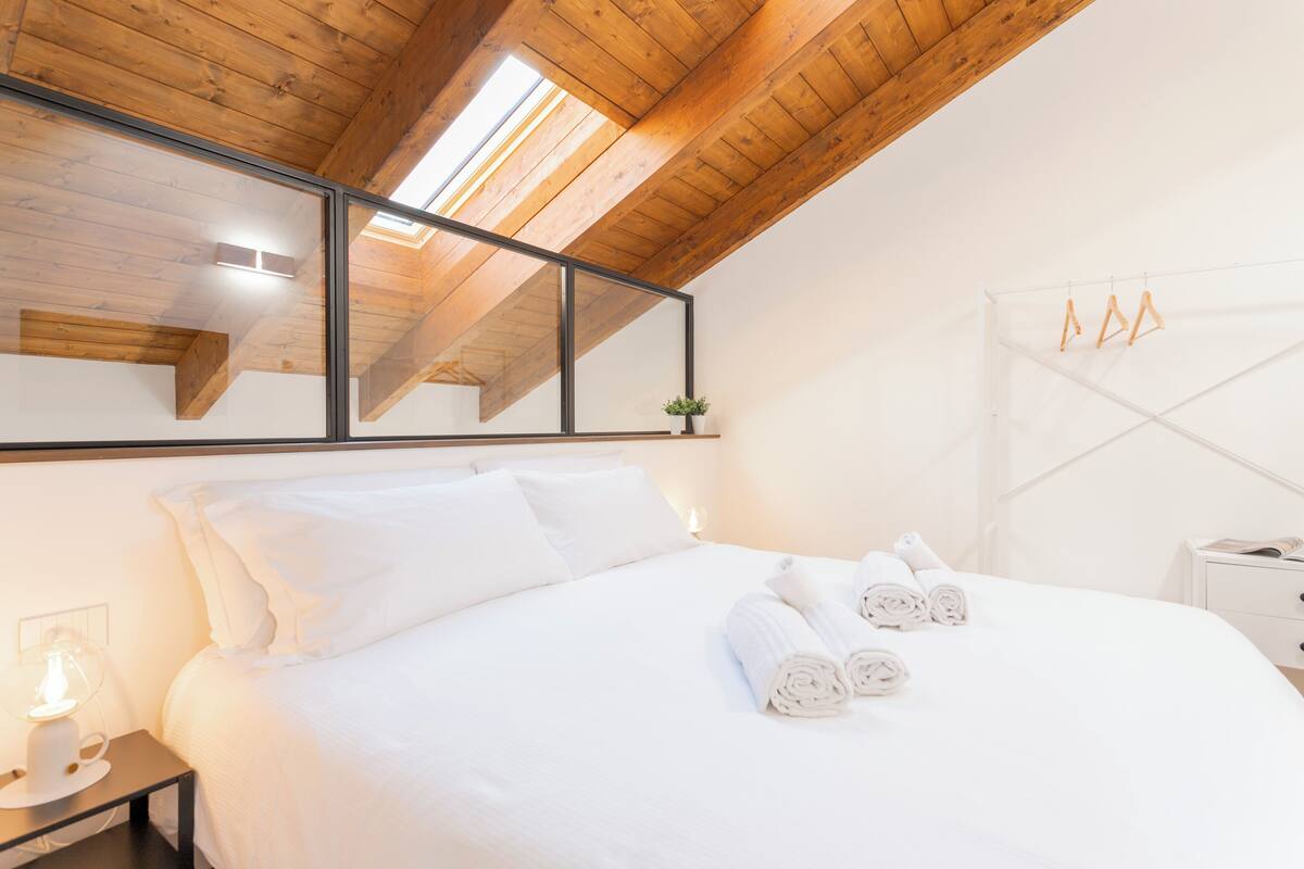 A hotel room with a wooden ceiling, a bed with white linens, a bedside table with a lamp, and a hanging clothes rack.
