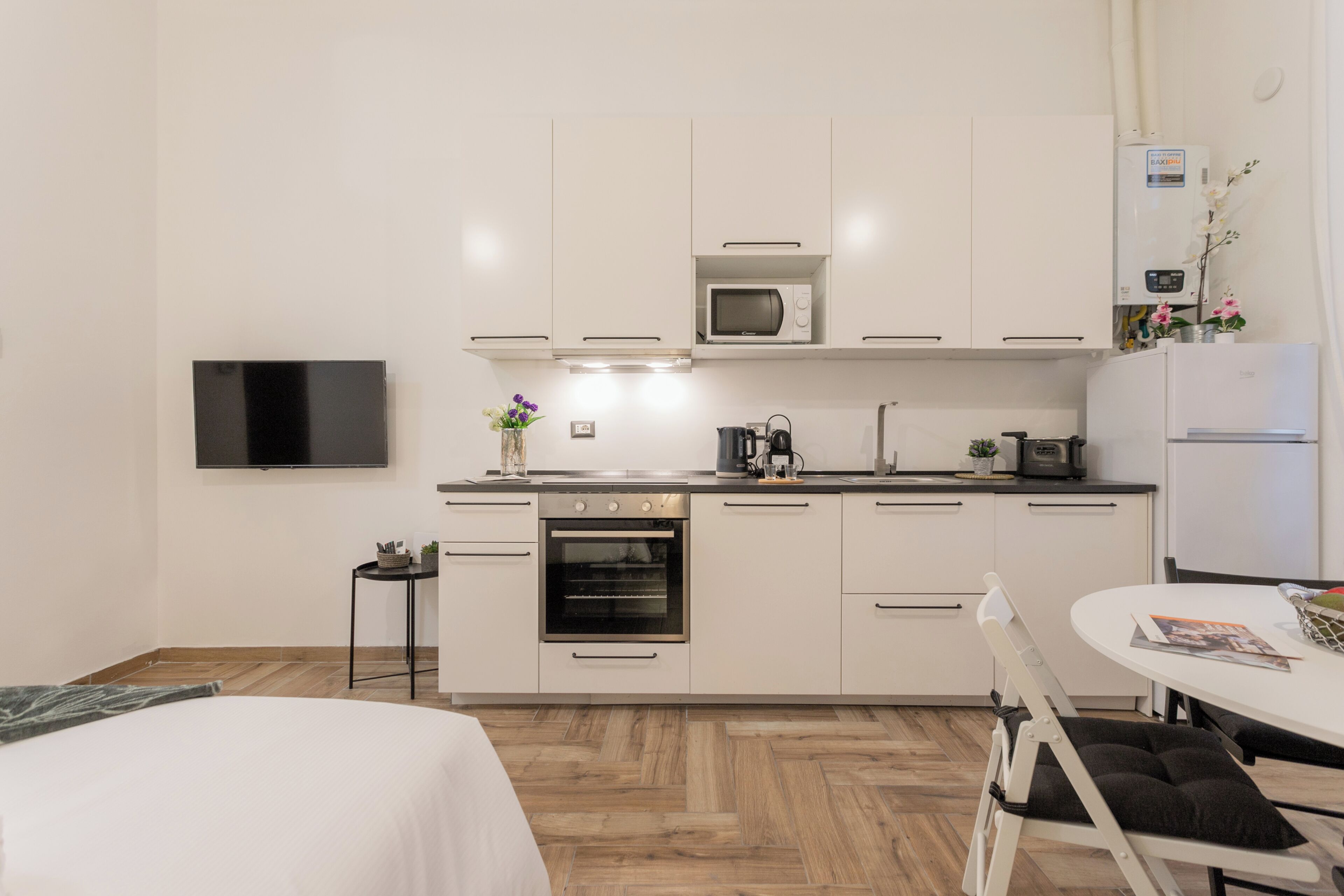 A modern kitchen with white cabinets, a microwave, an oven, and a refrigerator. There is a dining table with chairs and a flat-screen TV mounted on the wall.