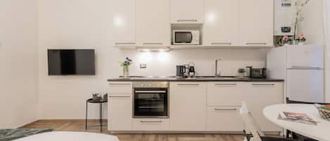 A modern kitchen with white cabinets, a microwave, an oven, and a refrigerator. There is a dining table with chairs and a flat-screen TV mounted on the wall.