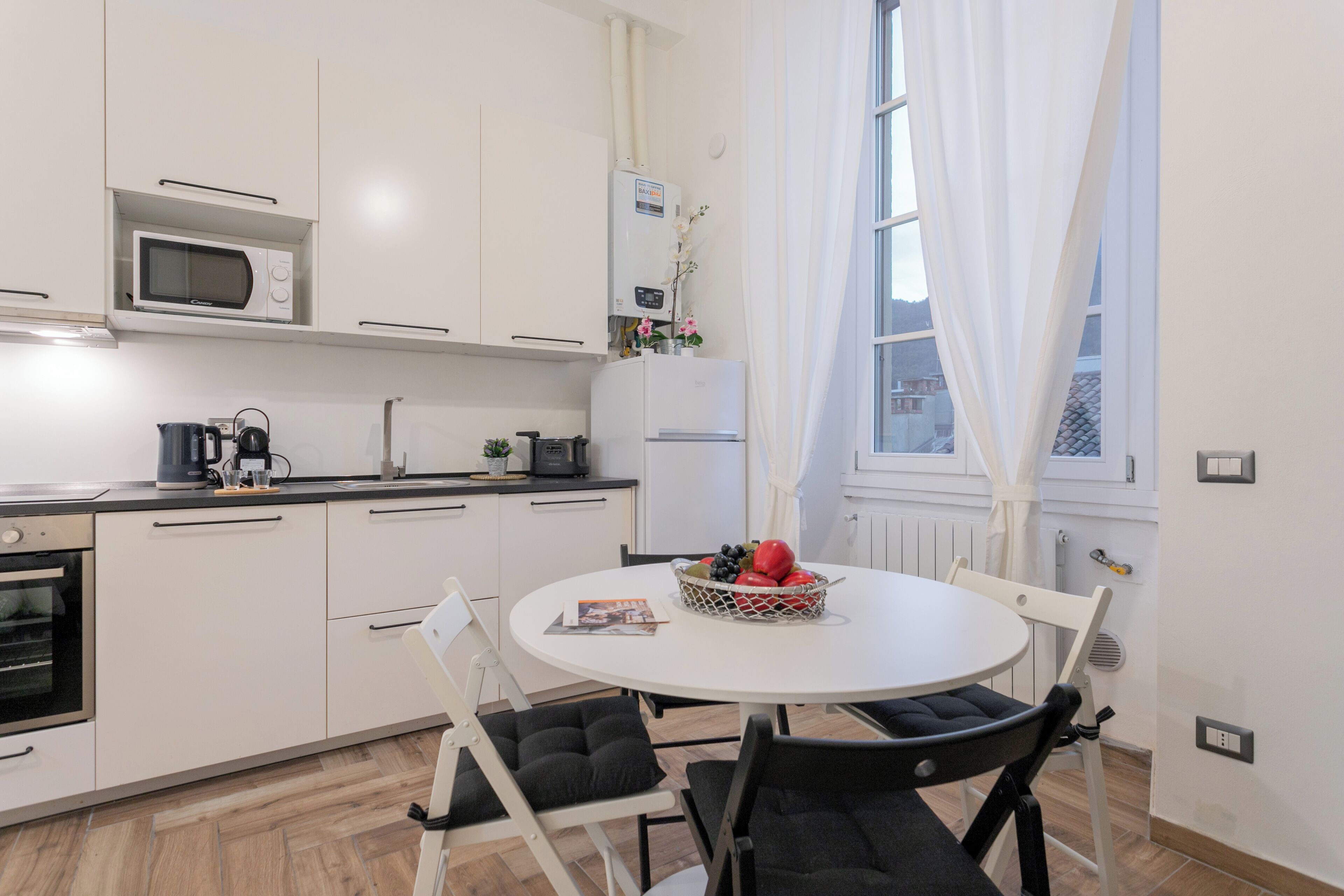 A modern kitchen with white cabinets, a microwave, coffee maker, and a refrigerator. There is a dining table with chairs and a fruit basket.