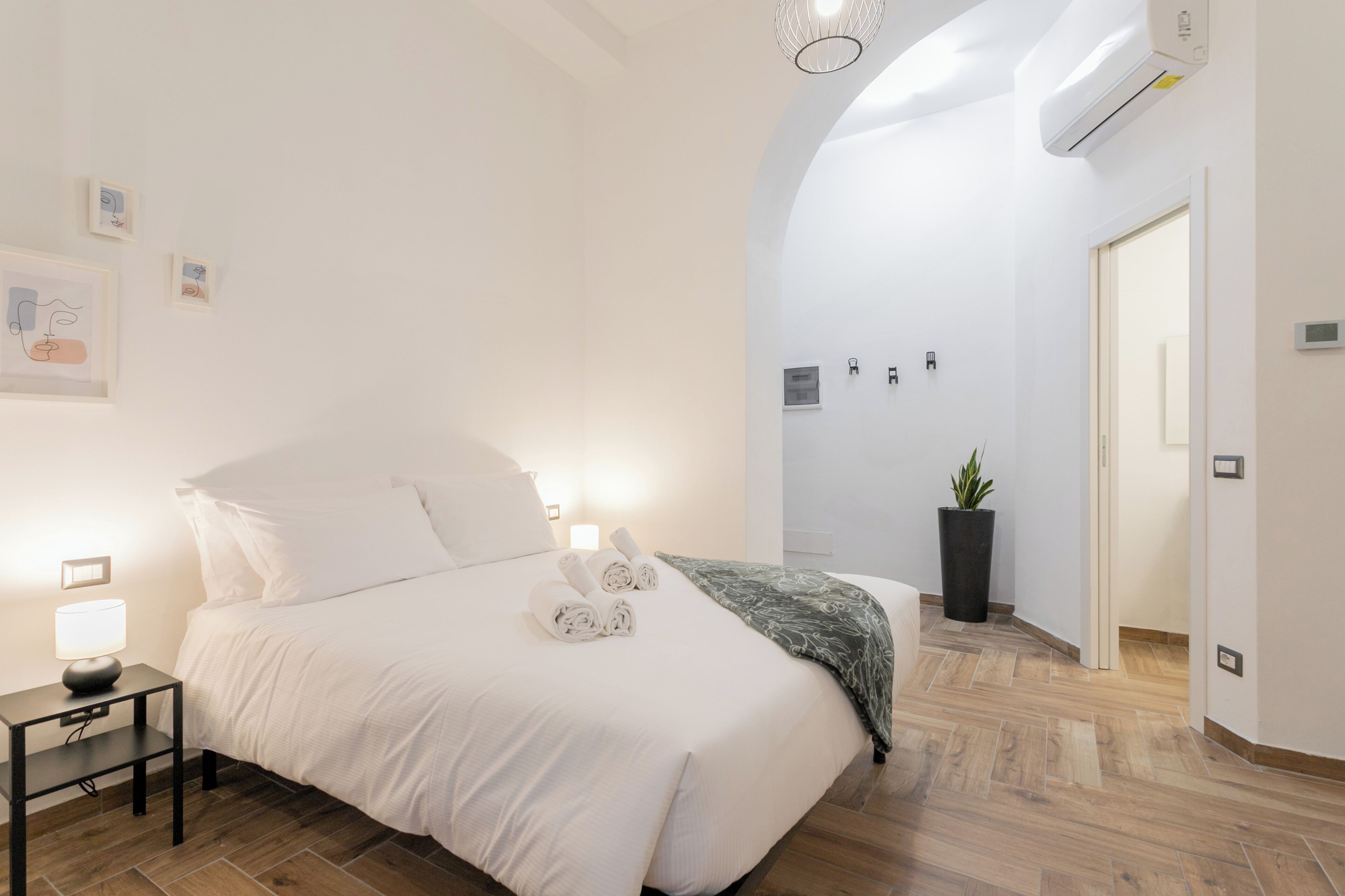 A modern hotel room with a neatly made bed, a bedside lamp, a potted plant, and a hallway leading to another room.