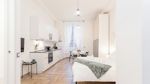 A modern kitchen with white cabinets, a dining area with a table and chairs, and a bedroom with a bed and a large window. - VOLTA COLLECTION BY HALLDIS (Como)