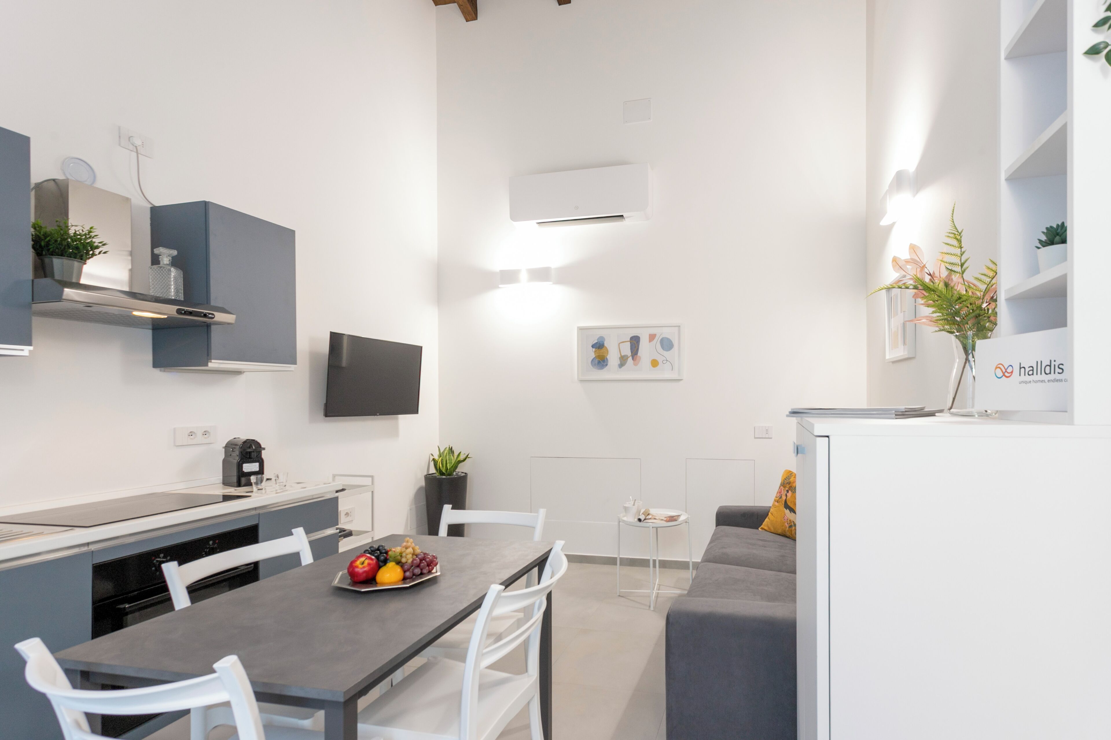 Une cuisine moderne avec une table à manger, des chaises, un panier de fruits et un canapé gris.