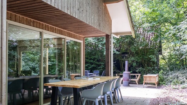 Una veranda esterna con tavolo e sedie di legno, circondata da vetrate e affaccio su una foresta.