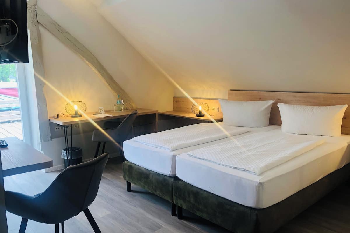 A hotel room with a loft-style ceiling, a desk with a chair, two beds with white linens, and a nightstand with a lamp.