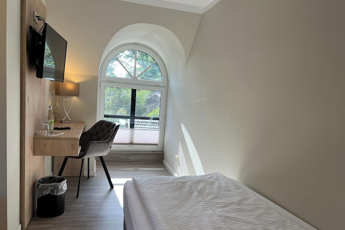 A bedroom with a bed, a desk with a chair, a TV, a window, and a trash bin.