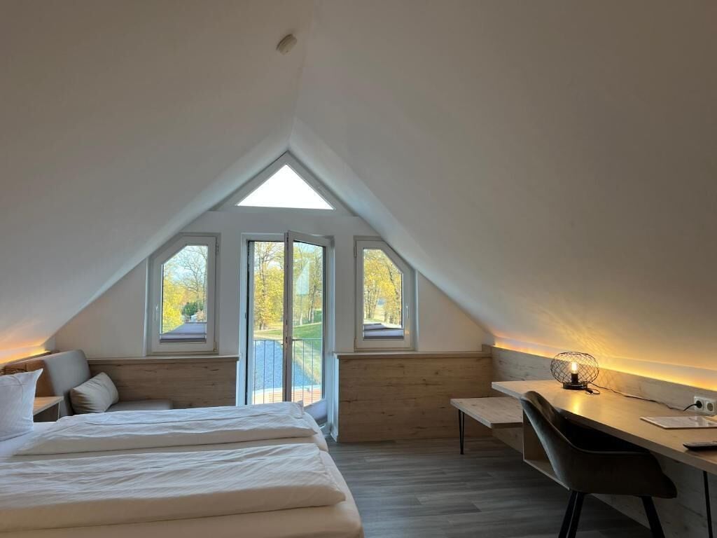 A bedroom with a bed, a desk, and a chair. There are three windows providing natural light.