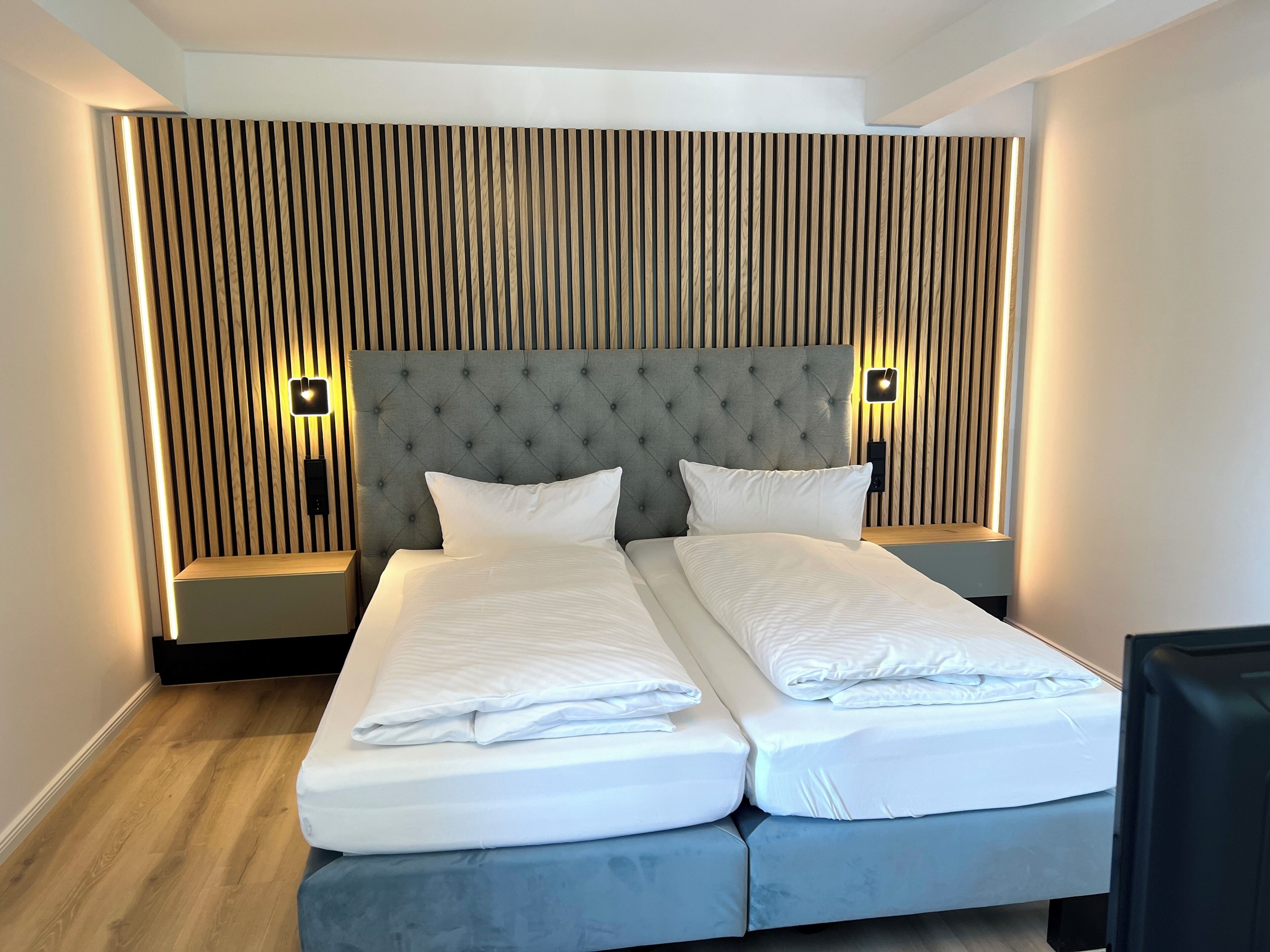 Una cama doble con cabecera capitonada, dos mesitas de noche y una pared con listones de madera verticales.