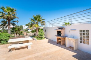 An outdoor patio with a stone barbecue and a sink, surrounded by palm trees and a white wall. - Villa Voramar with sea views. Cala D'Or (Cala d´Or)
