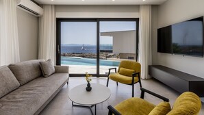 Een moderne woonkamer met een bank, gele fauteuils, een salontafel en een televisie. Een groot raam biedt uitzicht op een zwembad en de zee.