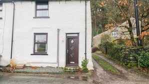 A white building with a brown door and windows, surrounded by trees and a stone wall. - Bobtail Cottage (Kendal)