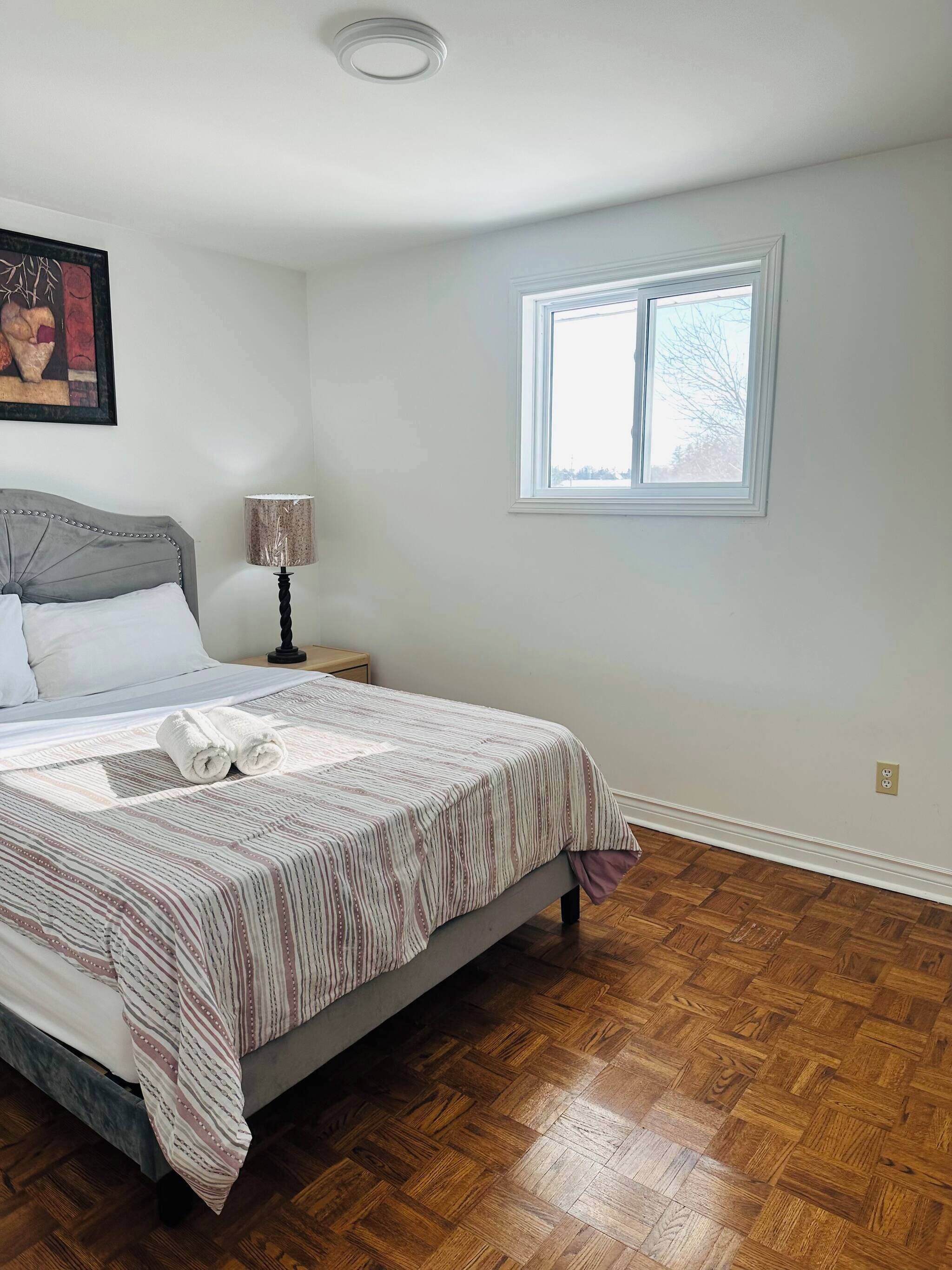 A bedroom with a bed, a nightstand, a lamp, a window, and a painting on the wall.