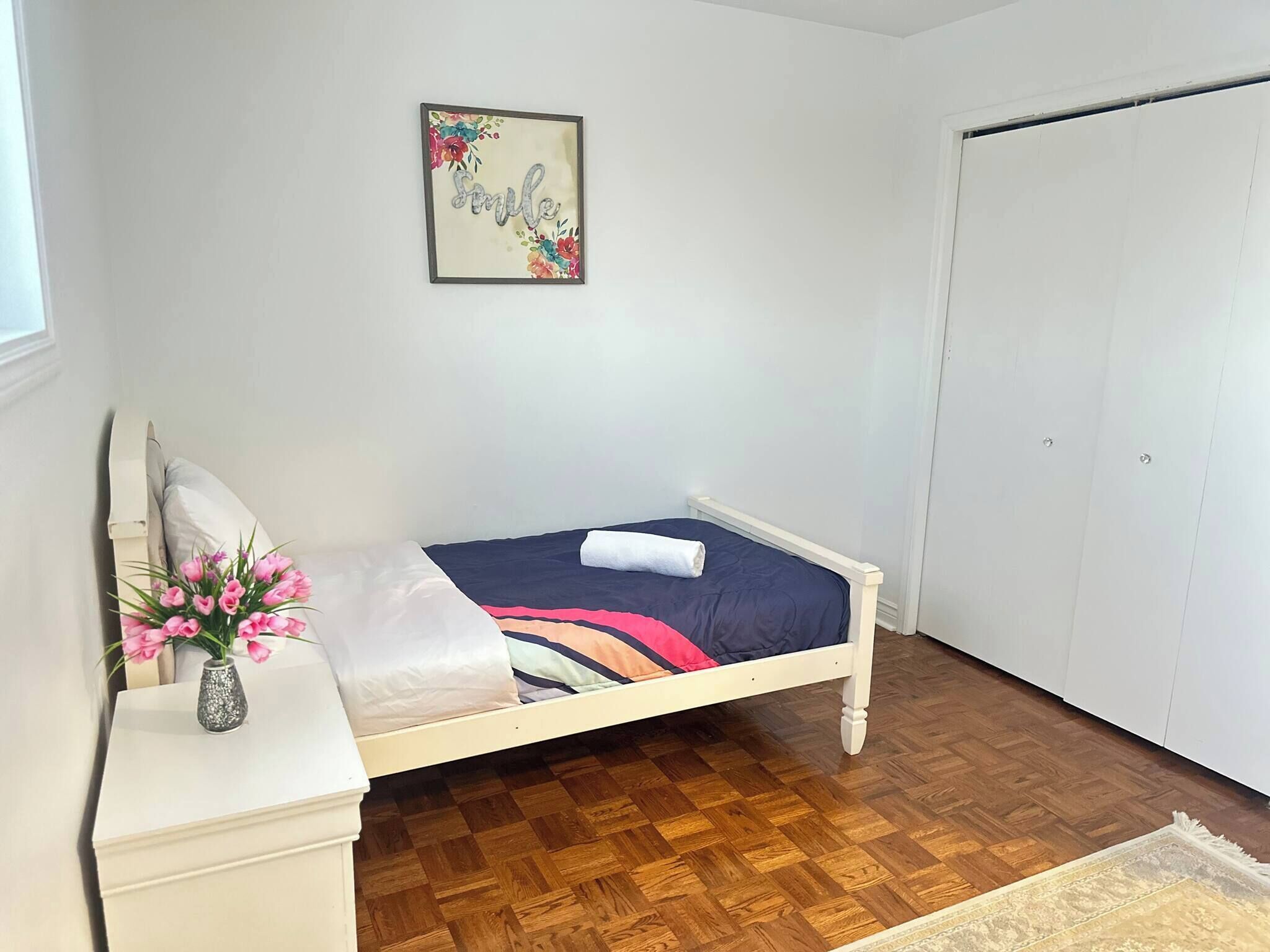 A bedroom with a bed, a bedside table with a vase of flowers, a wall-mounted picture, and a wardrobe.