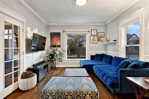 A living room with a blue sofa, a patterned ottoman, a television, and a window with a view of a house. - Remodeled Craftsman in Central District w/ Parking (Seattle)