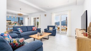 A modern living room with a sofa, armchairs, and a coffee table. There is a kitchen area with blue cabinets and a dining table. A TV is mounted on the wall, and there is a view of the outdoors through large windows. - Luxury Villa Blue Dream (Đenovići)