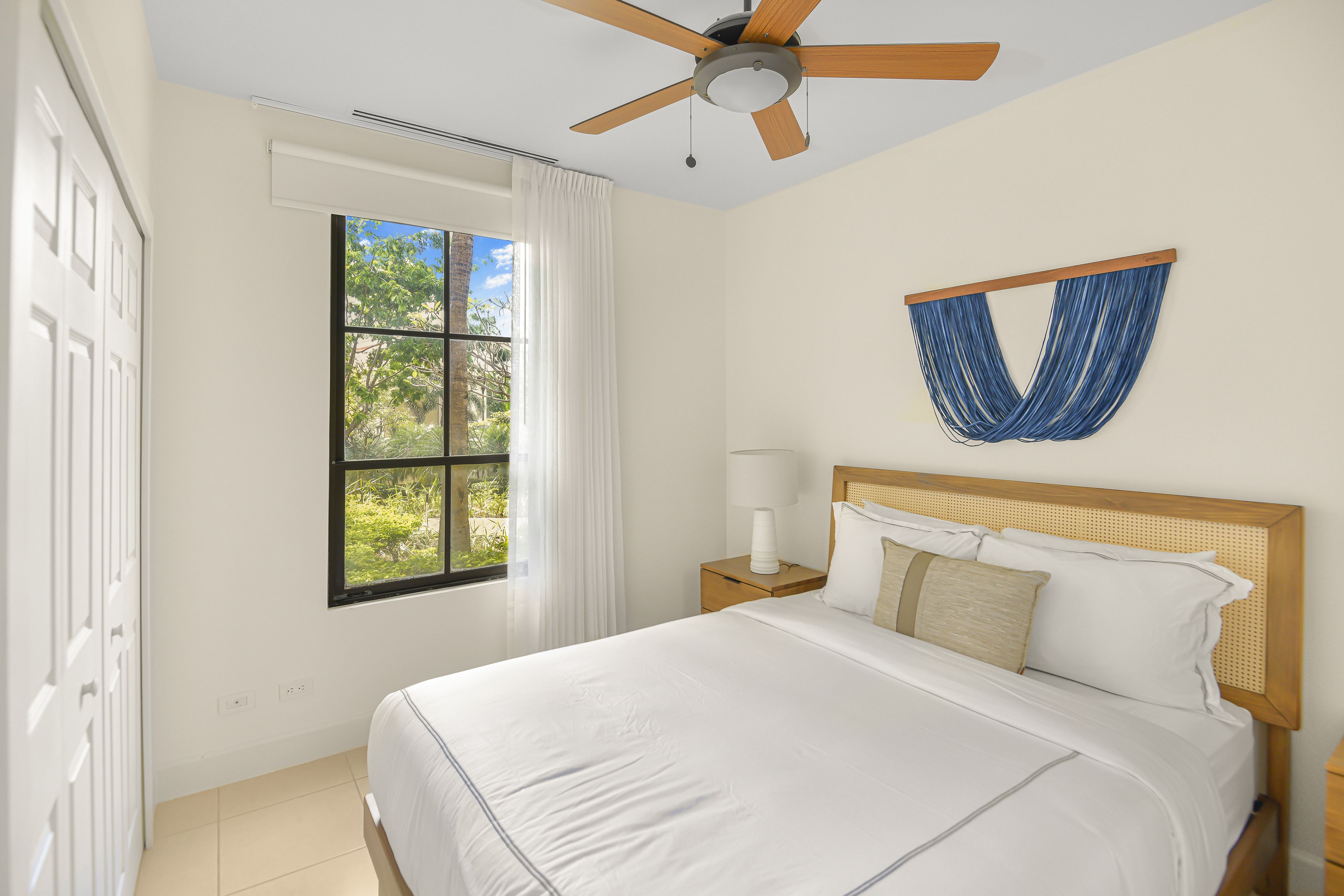 A bedroom with a bed, a nightstand, a lamp, a ceiling fan, and a window with curtains.
