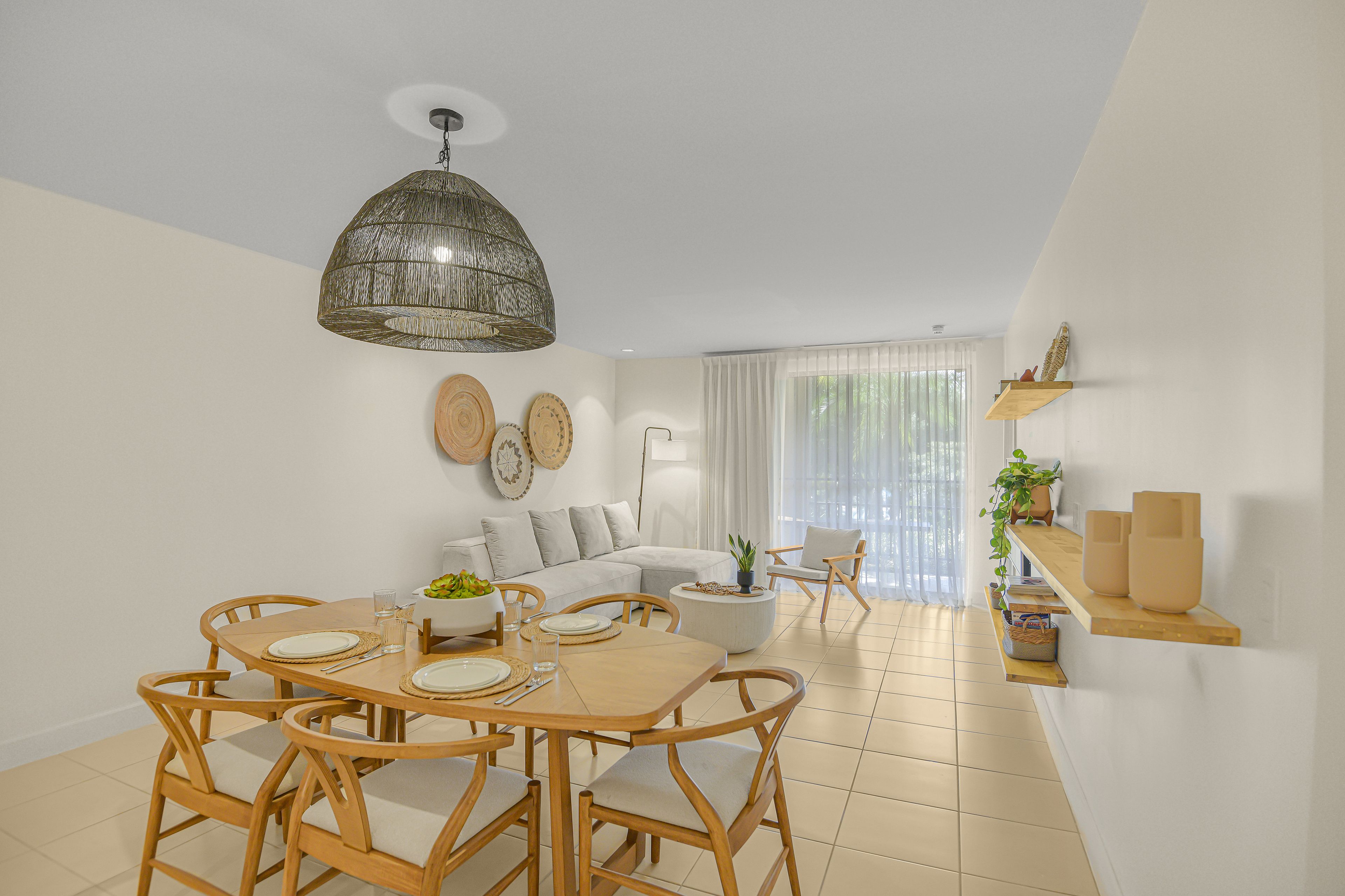 A modern living room with a dining area, featuring a sofa, a dining table with chairs, a hanging light fixture, and a large window with sheer curtains.
