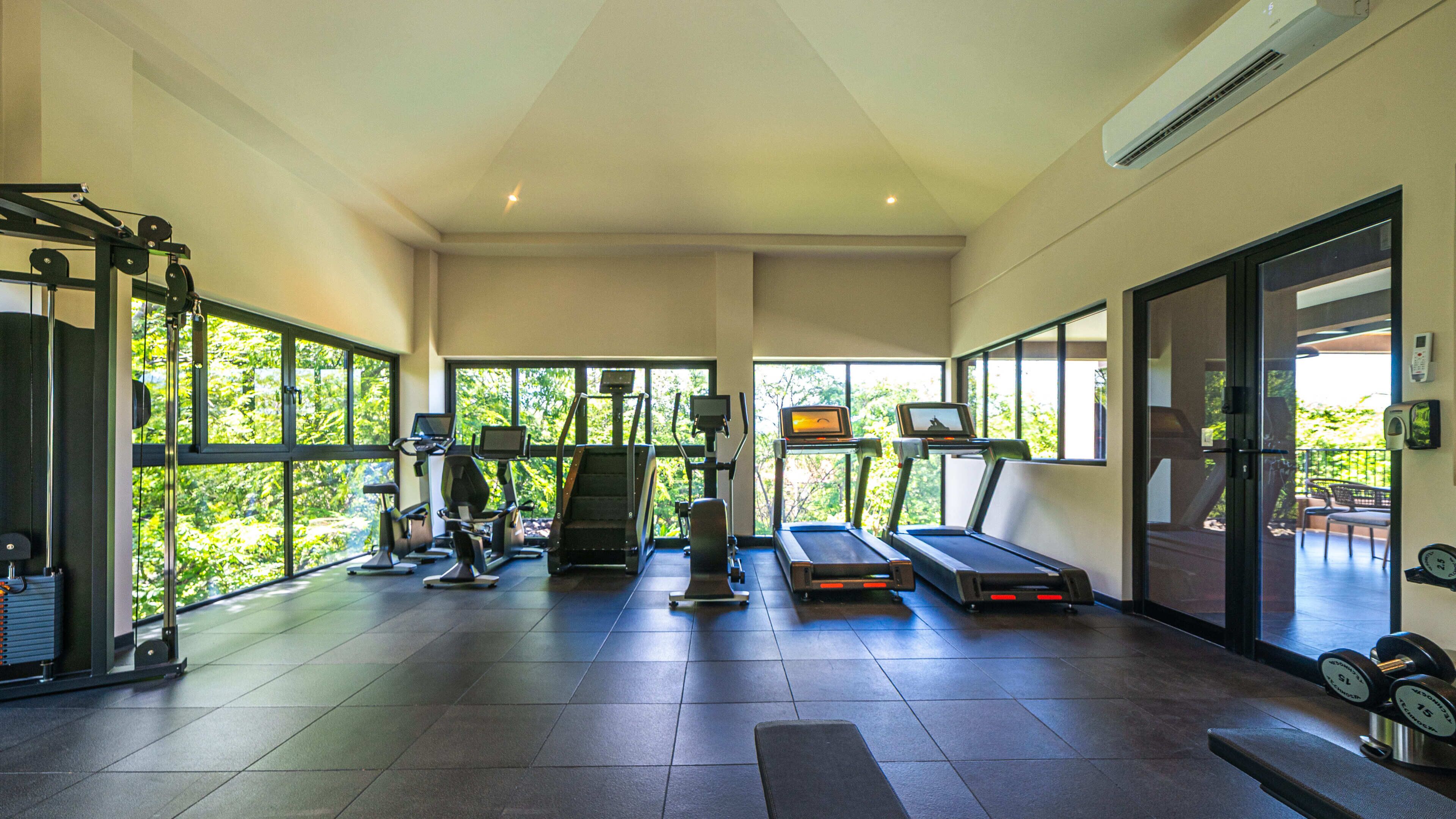 A spacious gym with various exercise equipment, large windows, and a view of greenery outside.