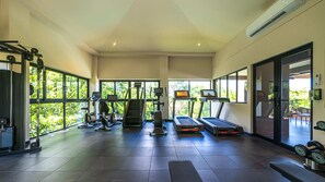 A spacious gym with various exercise equipment, large windows, and a view of greenery outside. - Pacifico Rentals (Sardinal)