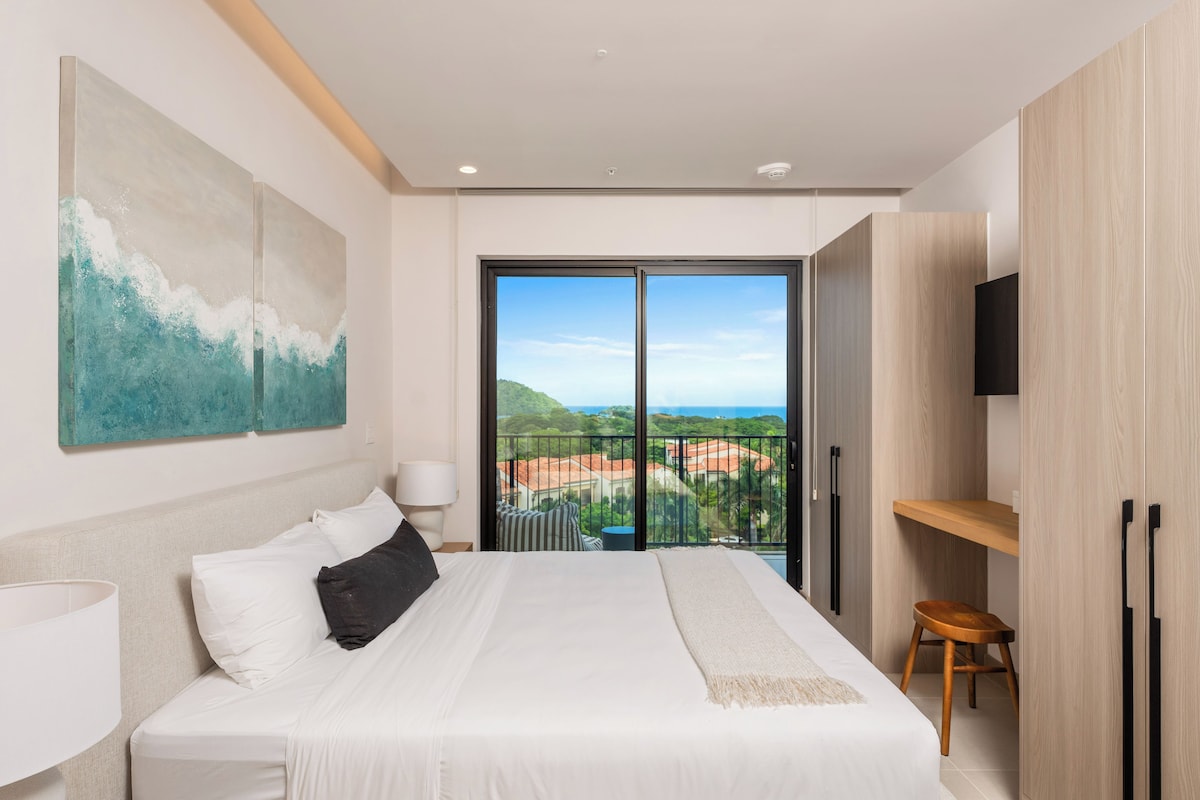 A bedroom with a large bed, a TV, and a balcony with a view.