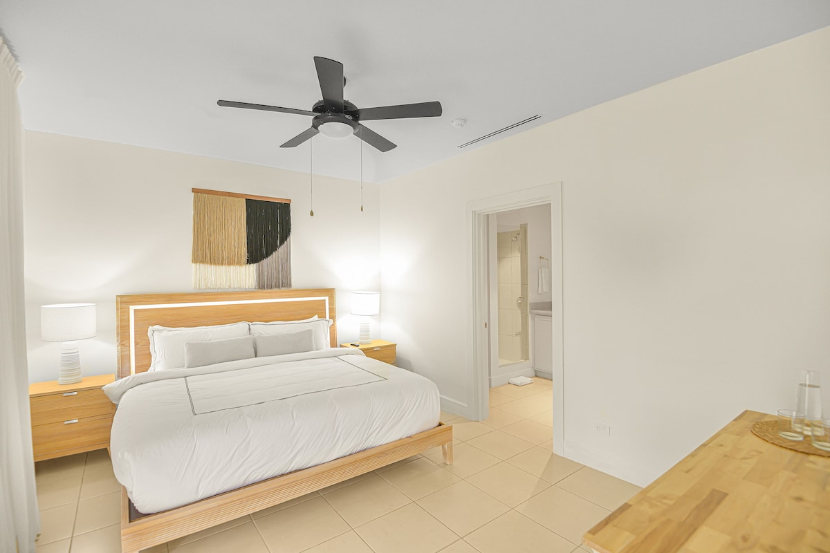A hotel room with a bed, bedside tables, a ceiling fan, and a door leading to a bathroom.