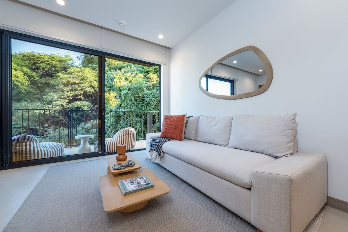 A modern living room with a sofa, a coffee table, and a large window offering a view of greenery.