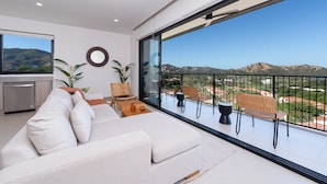 A modern living room with a large sofa, a dining table, and chairs, a kitchen area, and a balcony with a view of the mountains. - Pacifico Rentals (Sardinal)