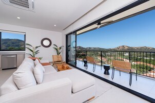 Una sala de estar moderna con un sofá grande, una mesa de comedor y sillas, una zona de cocina y un balcón con vistas a las montañas.