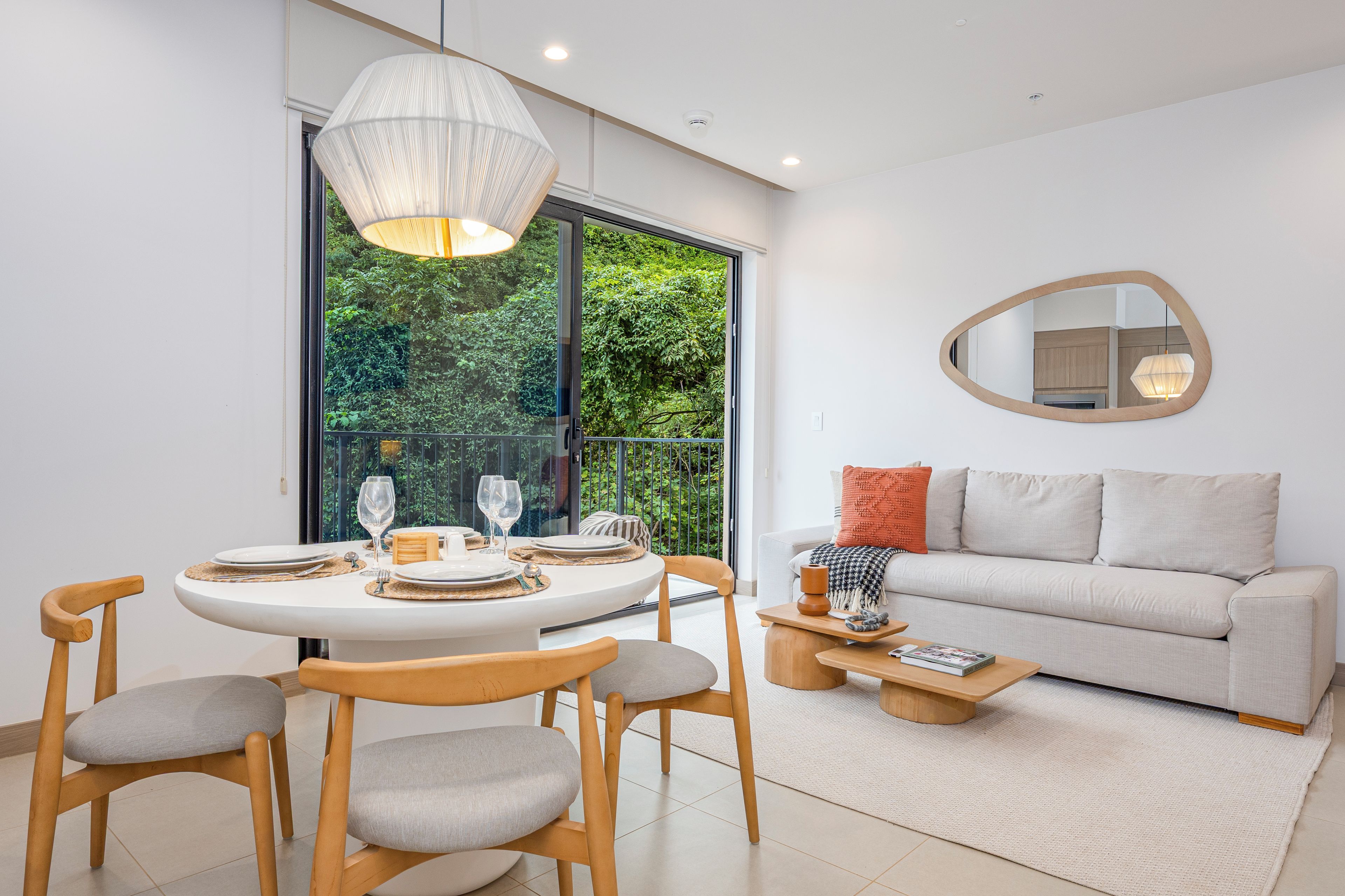 A modern living room with a dining area, a sofa, a coffee table, and a large window with a view of greenery.