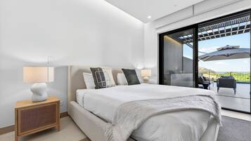 A modern bedroom with a large bed, a nightstand, a lamp, and a view through a glass door to an outdoor patio.