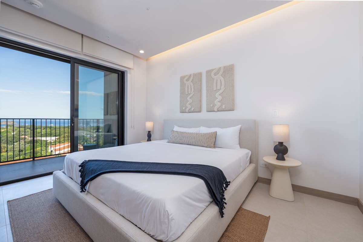 A modern bedroom with a large bed, a nightstand, a lamp, and a view of the outdoors.