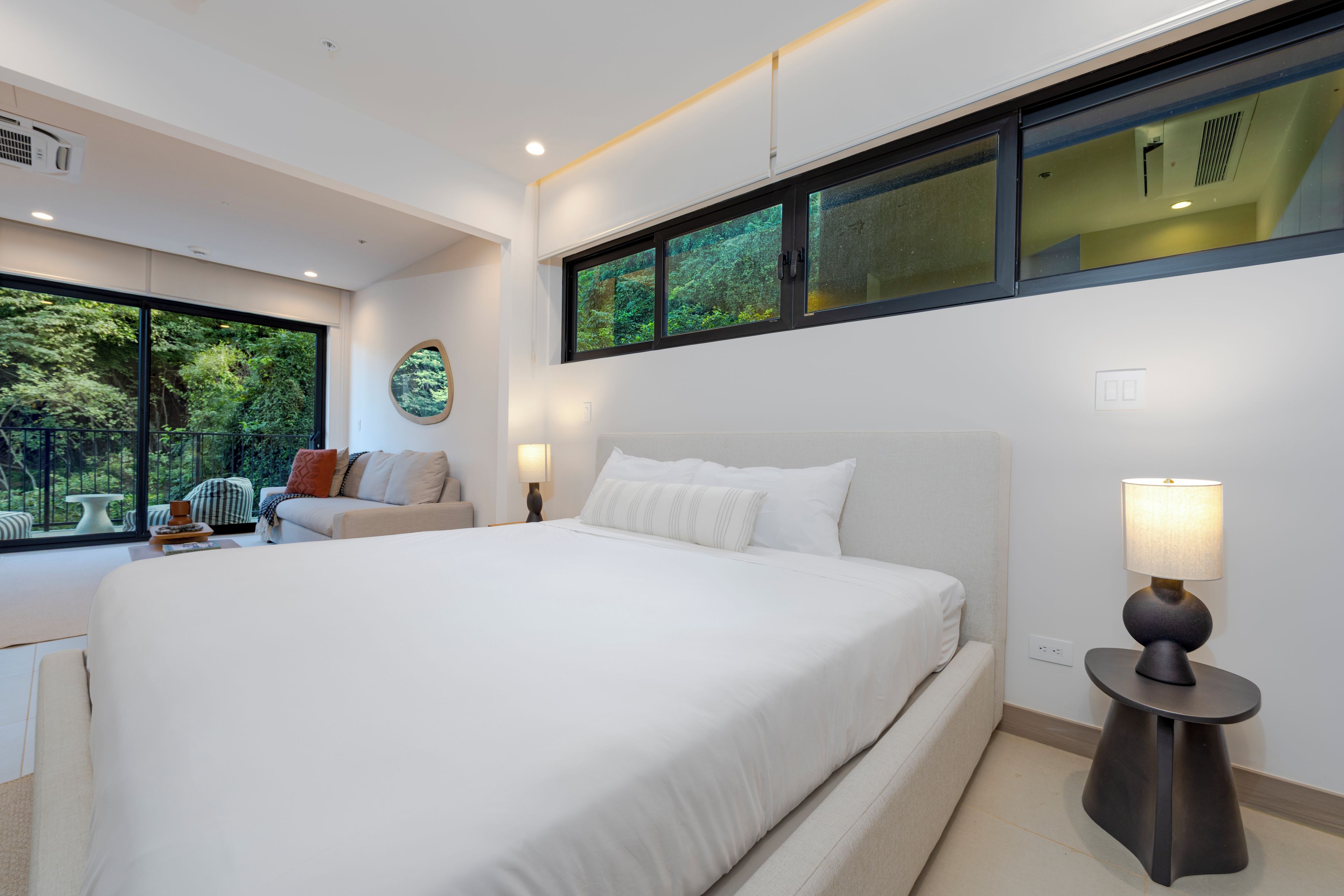 A modern bedroom with a large bed, a bedside table, a lamp, and a view of greenery through a window.