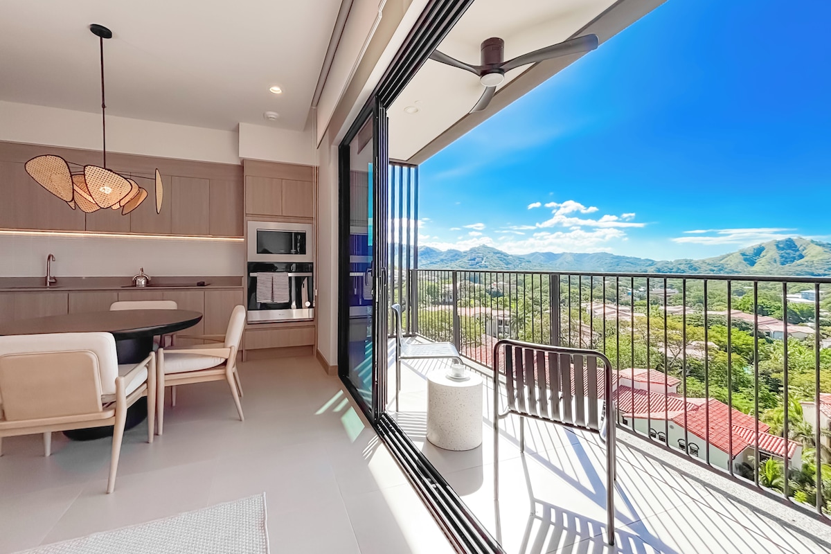 A modern kitchen with a dining area, a balcony with a view of the mountains, and a ceiling fan.
