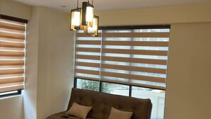 A living room with a brown sofa, two chairs, a dining table, and large windows with blinds.