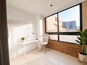 Una habitación moderna con un ventanal grande, un escritorio con silla, una planta en maceta y una pared de ladrillo.