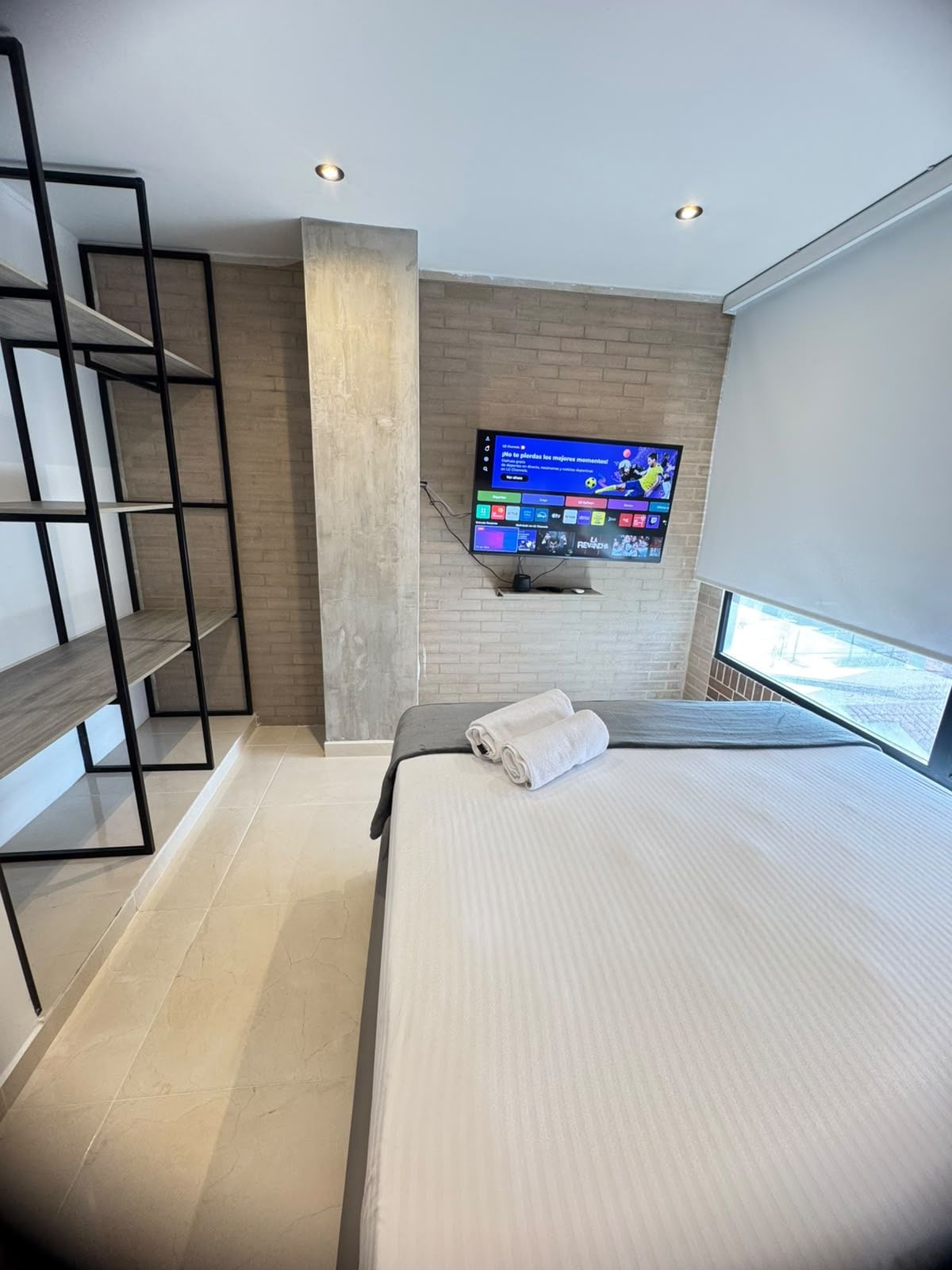 Une chambre d’hôtel moderne équipée d’un lit, d’un téléviseur fixé au mur, d’une étagère en bois et d’une fenêtre donnant sur un paysage.