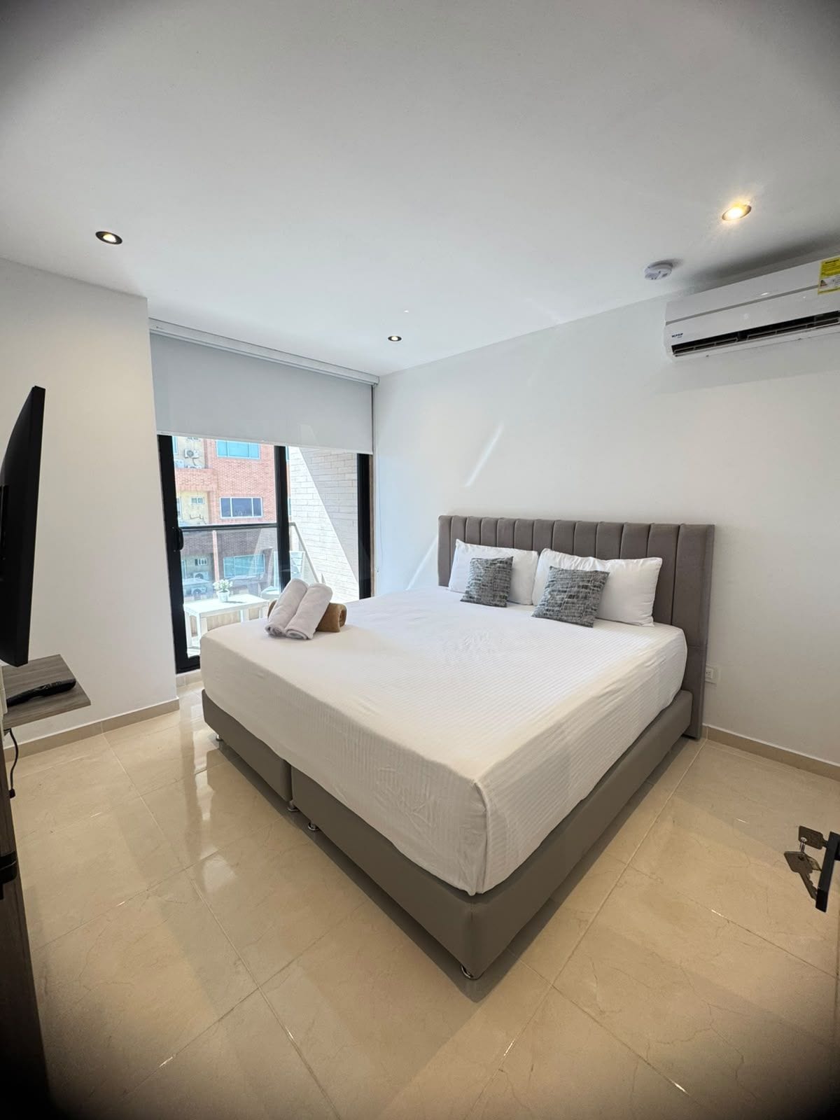A modern bedroom with a large bed, a flat-screen TV, and a window with a view of buildings.