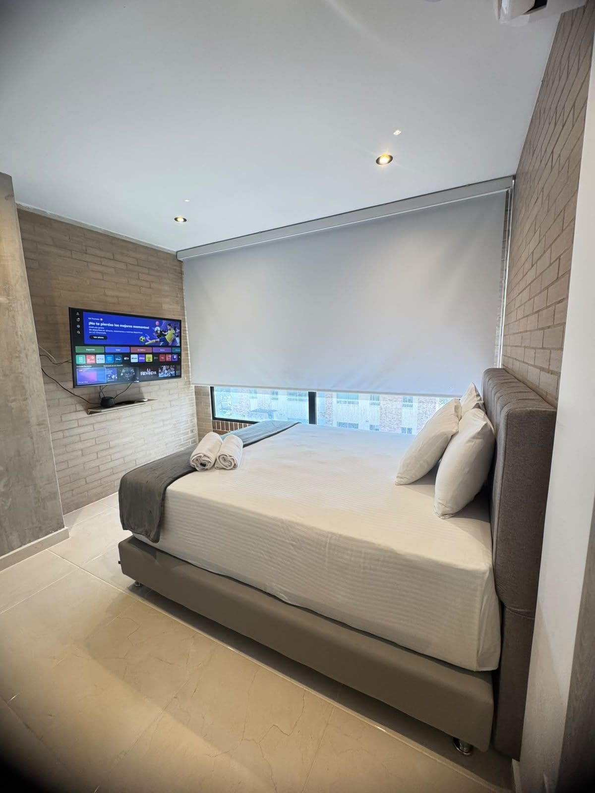 A modern bedroom with a large bed, a mounted TV, and a city view.