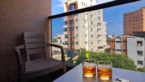 Een balkon met een bakstenen muur, een houten stoel, een tafel met twee glazen amberkleurige vloeistof, en een smartphone.