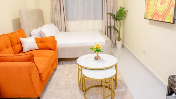 Une petite pièce bien éclairée, avec un lit, un canapé orange, une petite table ronde ornée d’un vase de fleurs et une plante en pot.