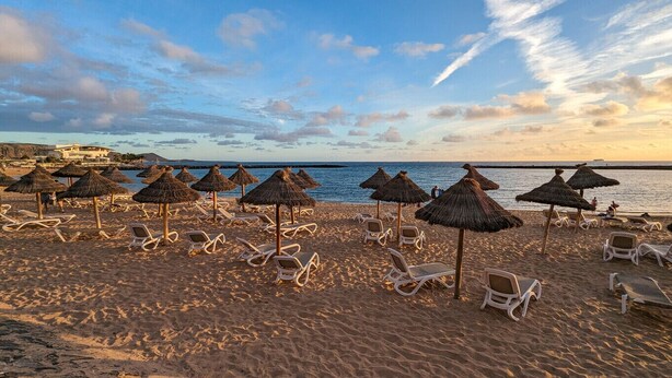 Ein Strand mit Liegen und Reetdachsonnenstühlen, ein klarer Himmel und ein Blick auf eine Küstenstadt in der Ferne.