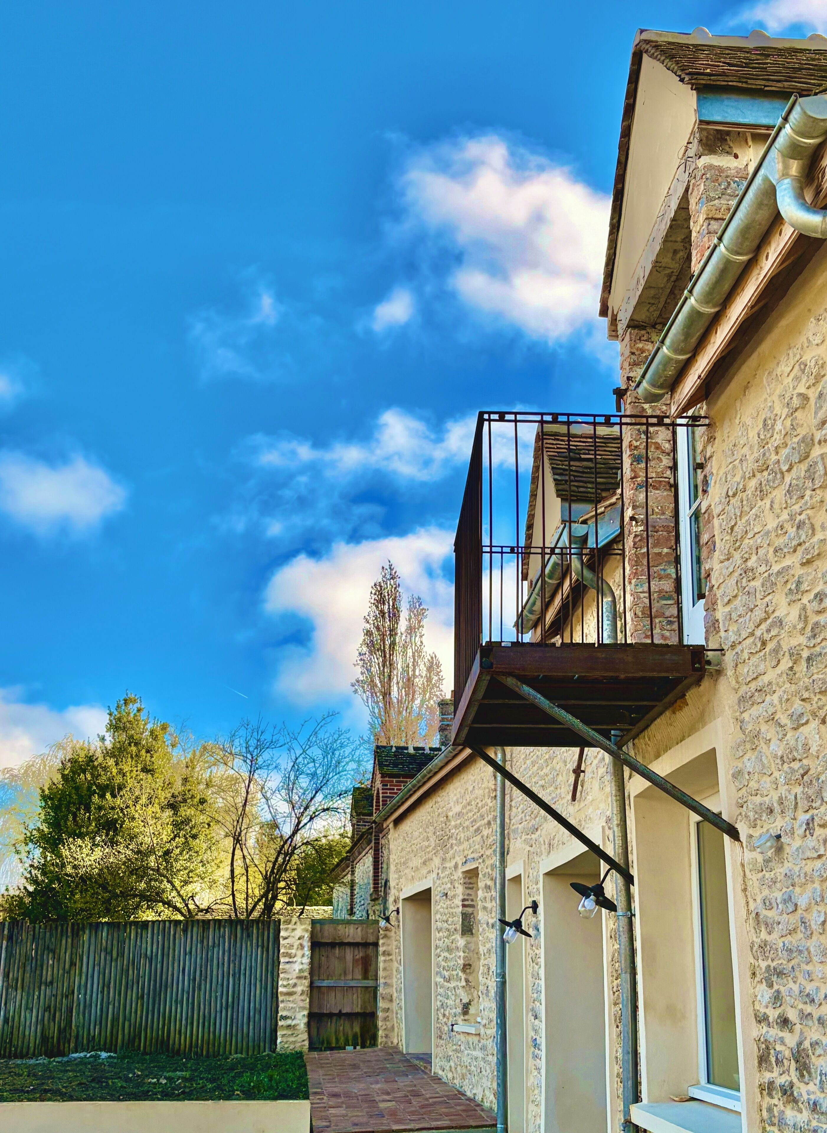 Un bâtiment en pierre doté d’un balcon, d’une clôture en bois et surplombant un ciel bleu dégagé.